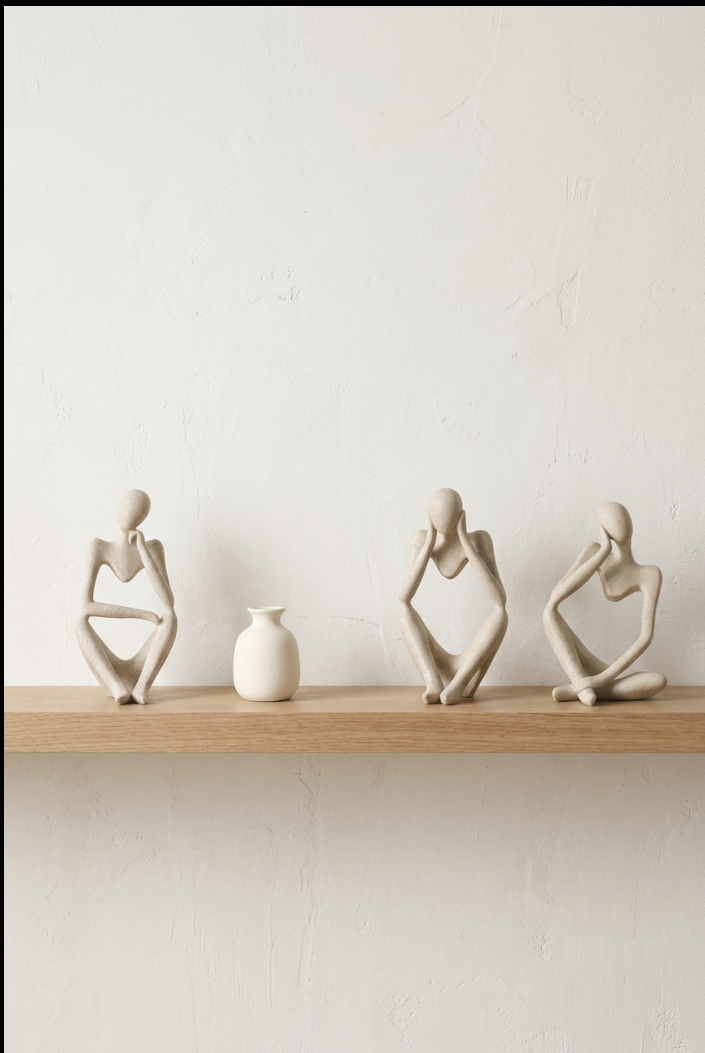 Three sand-toned abstract thinker figurines and a small white vase on a light oak shelf against a warm white wall