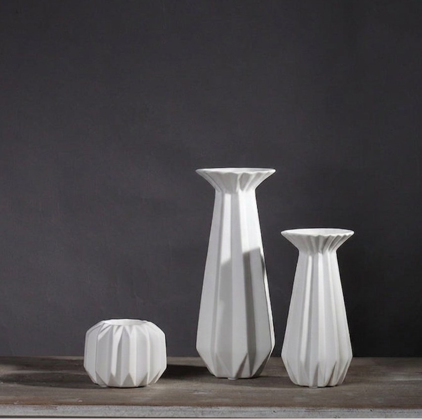 Three white faceted porcelain candle holders in graduated sizes — one tall, one medium and one small round — on a rustic wood surface against a dark gray wall.