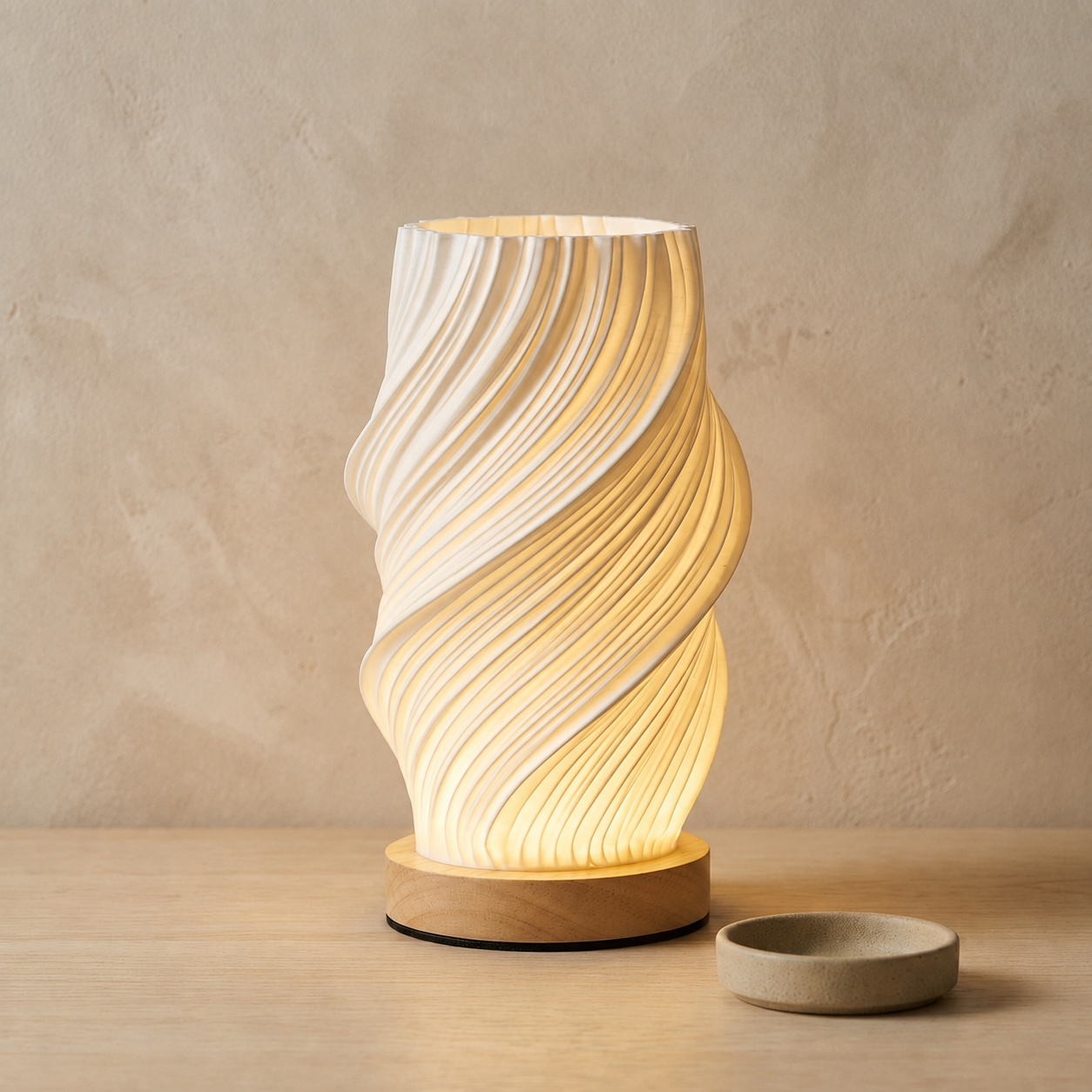 White spiral 3D-printed table lamp with a warm glow on a wooden base beside a small stoneware dish against a beige wall
